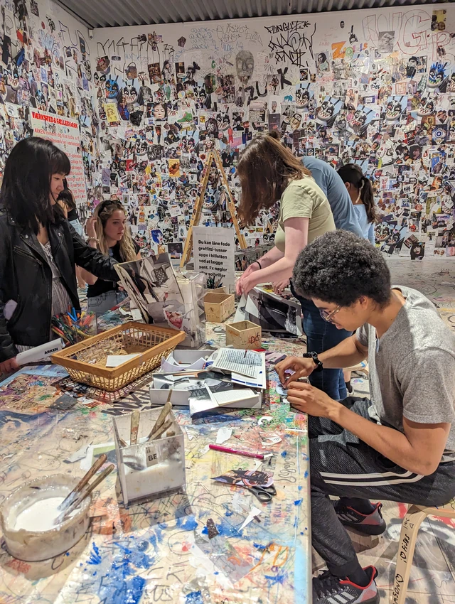 Students work with paper and small physical constructions around a shared table in a museum workshop covered with collage images.