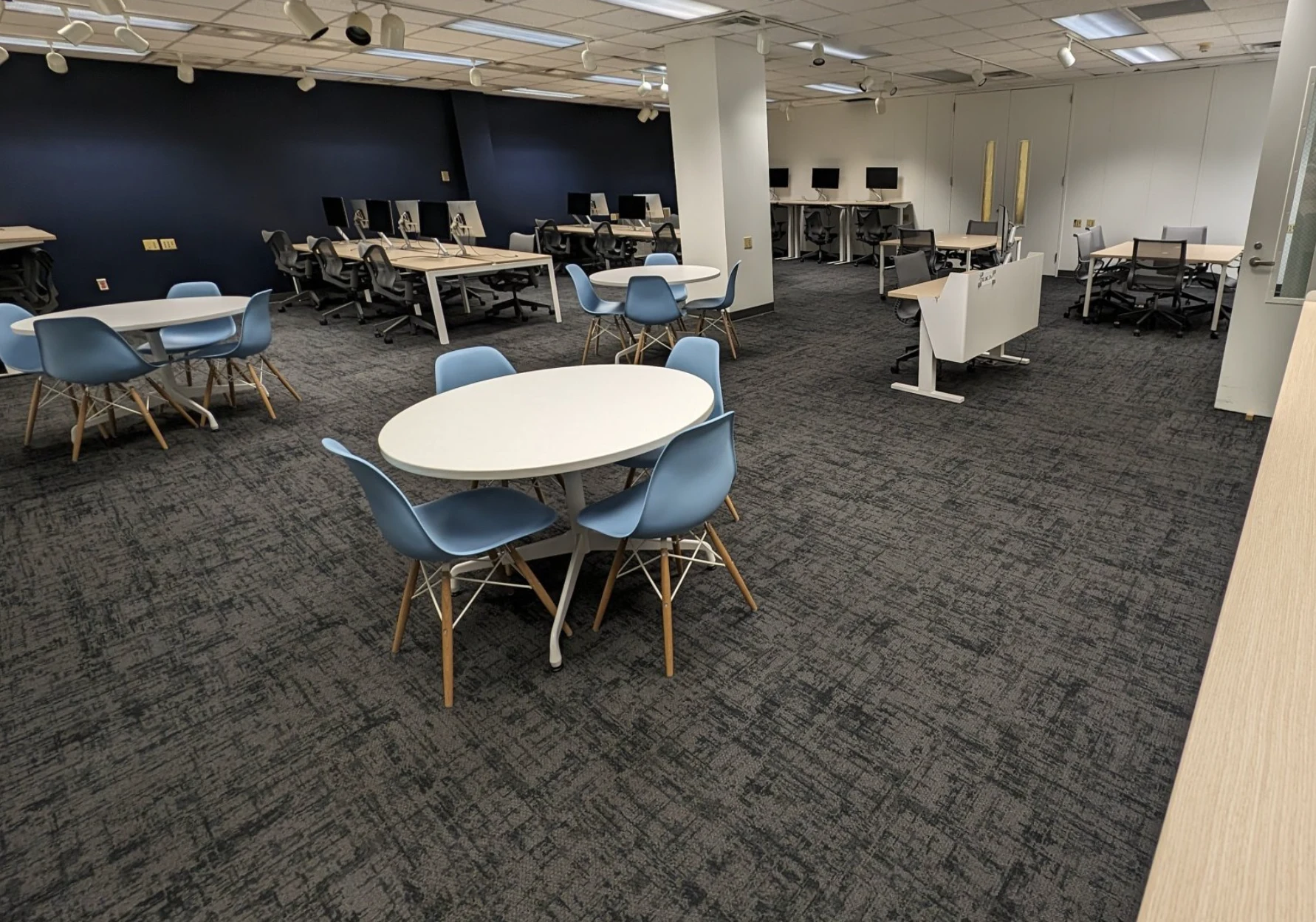 Photograph of the existing Co-Lab with white tables, blue chairs, and its original dark patterned carpet