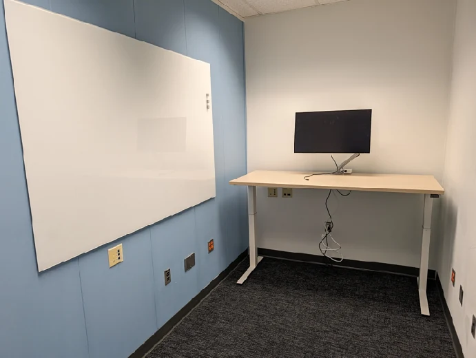 The small renovated Sitterson 144 room with a monitor, adjustable desk and wide wall-mounted writing board.