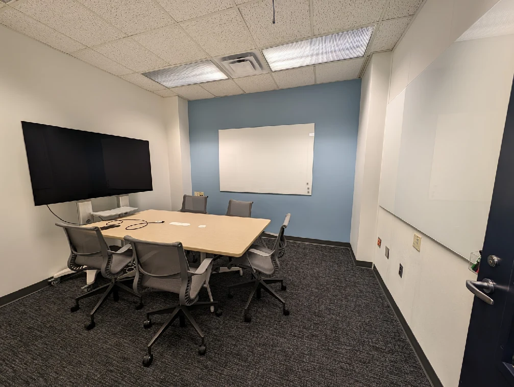 Sitterson 135 after renovation with a shared meeting table, a display, chairs and writing boards.