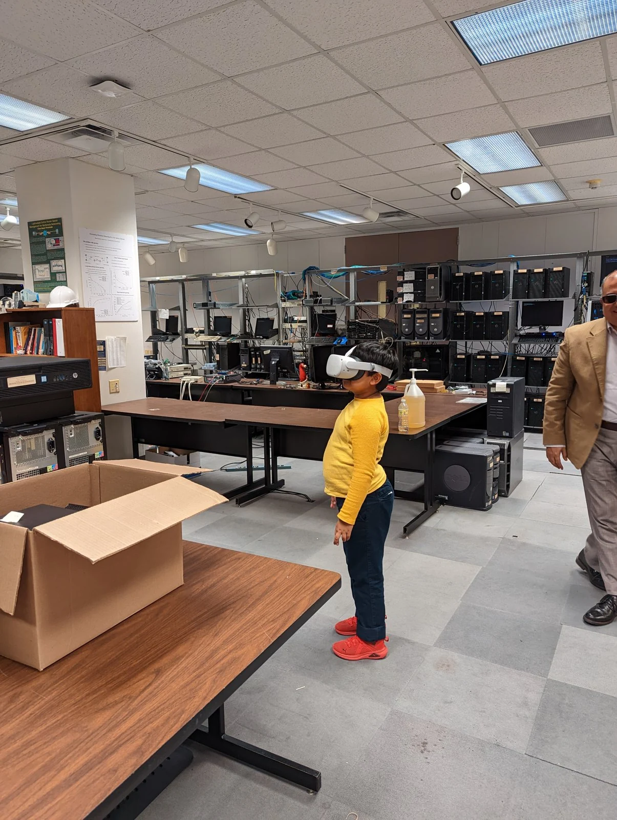 A child tries a white VR headset among the old lab tables and computer racks, with an adult at the right edge