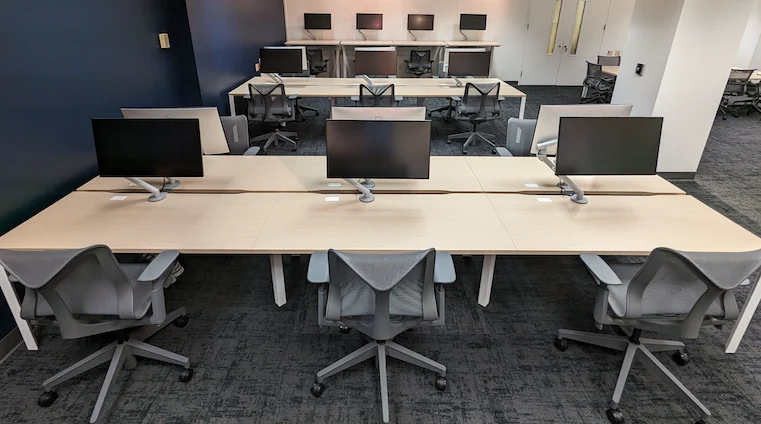 Installed white table bases and monitor arms with light wood surfaces and darker task chairs against a navy wall.