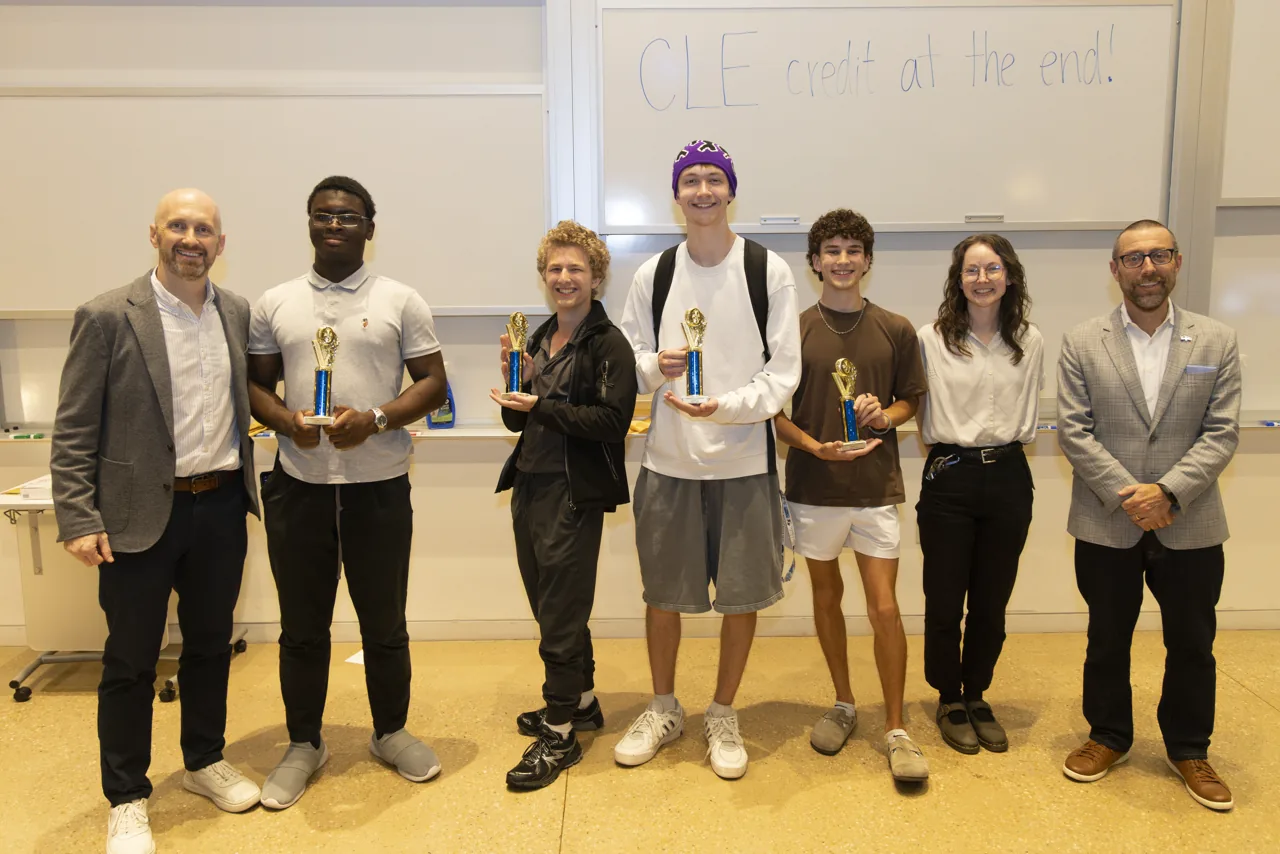 A group posing at the front of the room with several trophies